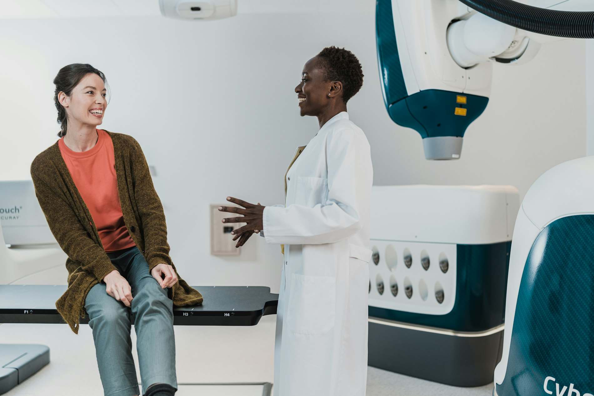 Médica de jaleco conversando com uma paciente sentada num consultório claro